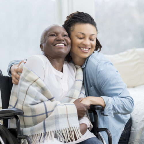 Young woman embraces her beautiful senior mother who is in a wheelchair. The adult daughter is visiting her mom in her mom's home.