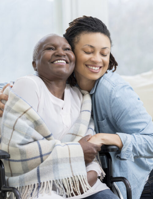 Young woman embraces her beautiful senior mother who is in a wheelchair. The adult daughter is visiting her mom in her mom's home.